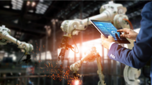 An image of a man in a suit holding a tablet, controlling manufacturing equipment in front of him